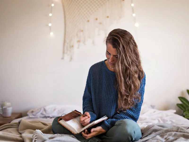 Woman in blue sweater sat on her bed writing in her leatherbound journal